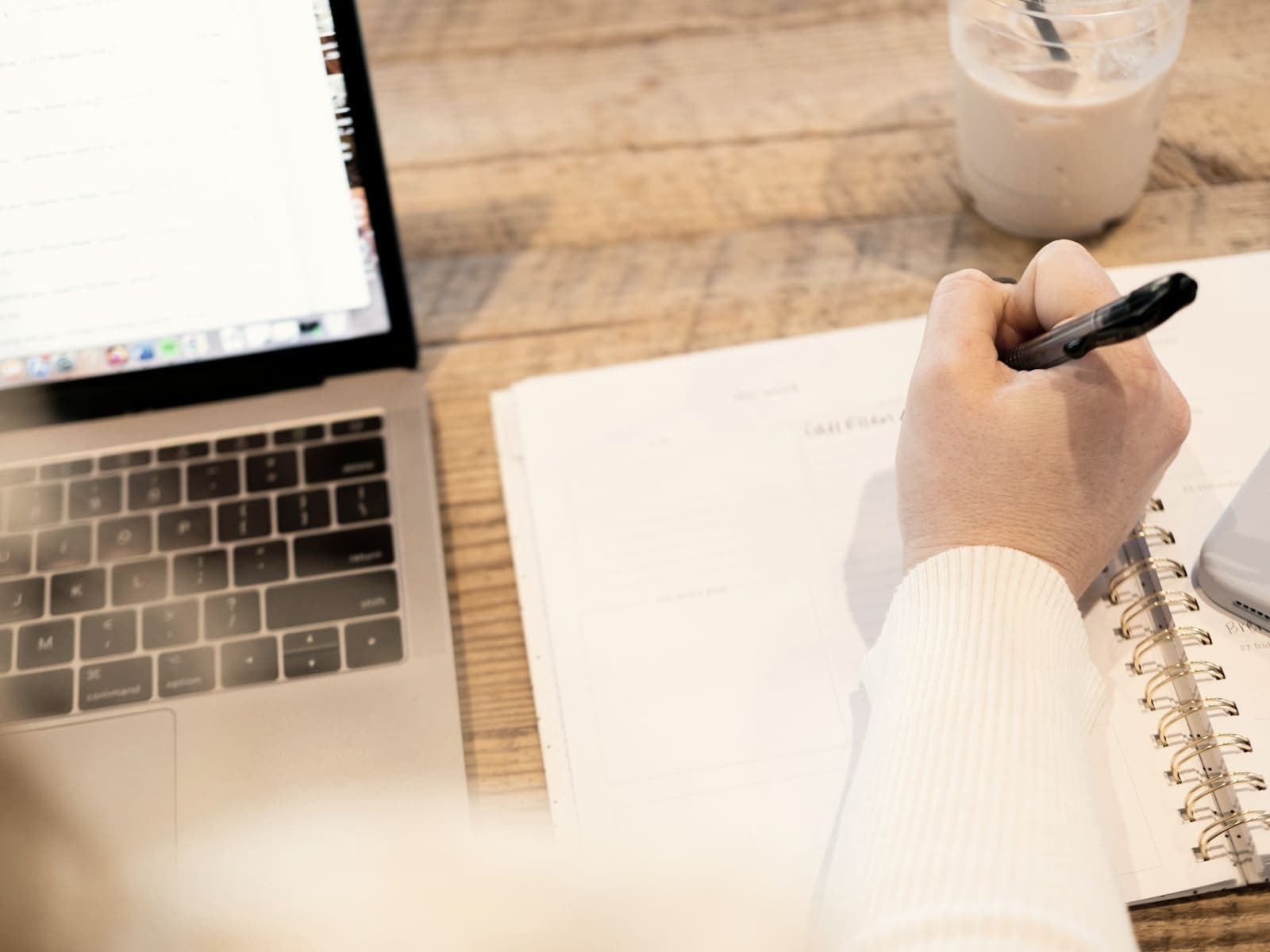 Someone writing notes in a notebook beside an open laptop